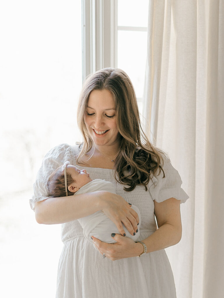 mom standing by window holding newborn baby girl